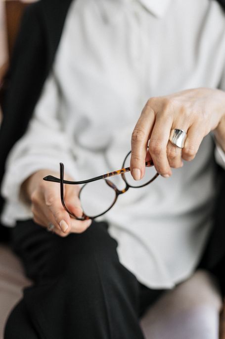 Person hält eine Brille mit dunklem Gestell vor sich, trägt weißes Hemd und schwarze Hose